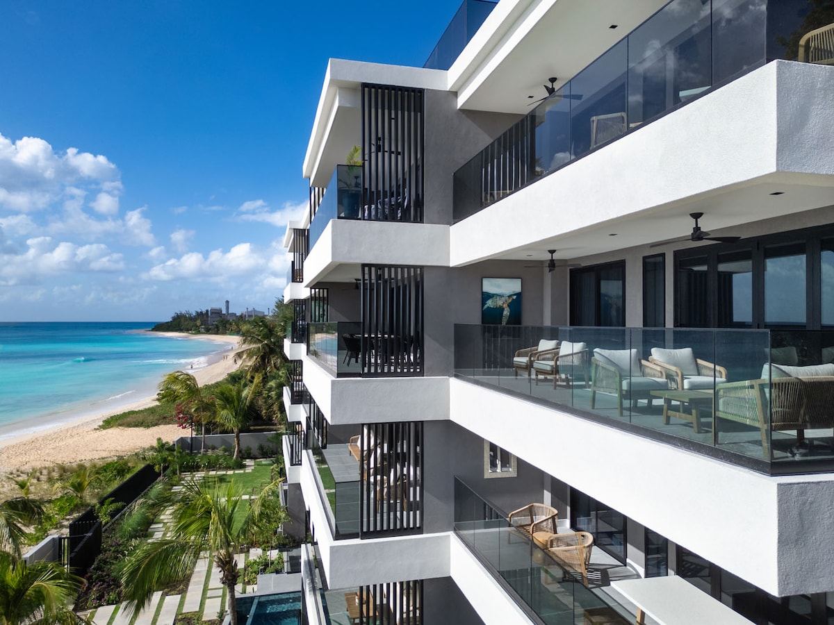 A modern beachfront condominium is displayed from an angle, featuring multiple balconies with glass railings. Each balcony showcases comfortable seating arrangements and offers stunning views of the sandy beach and turquoise ocean, framed by palm trees swaying gently in the breeze.
