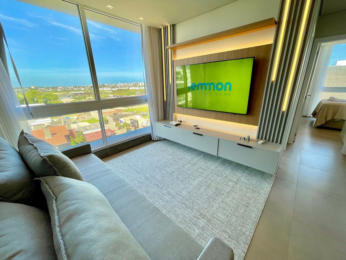 A modern living area is highlighted by a large window offering views of the surrounding landscape. A comfortable sofa faces a mounted television, while a soft area rug lays beneath. Natural light fills the space, accentuating the neutral color palette.
