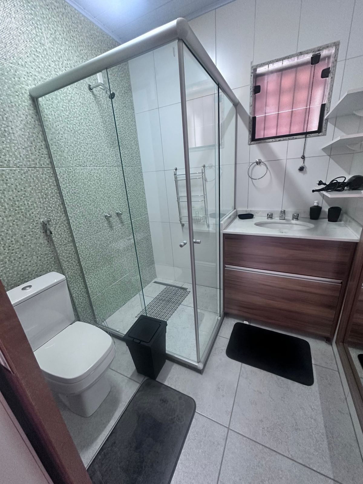 The bathroom is equipped with a glass-enclosed shower featuring modern fixtures. A compact toilet is situated beside a wooden vanity with ample storage. Neutral tiles and a decorative accent wall create a clean look, complemented by two bath mats on the floor.
