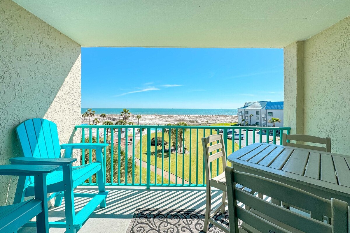 A corner balcony is presented, featuring a dining table and chairs alongside an adirondack chair. Expansive views of the beach and coastline extend beyond, showcasing the Gulf of Mexico under a clear blue sky.
