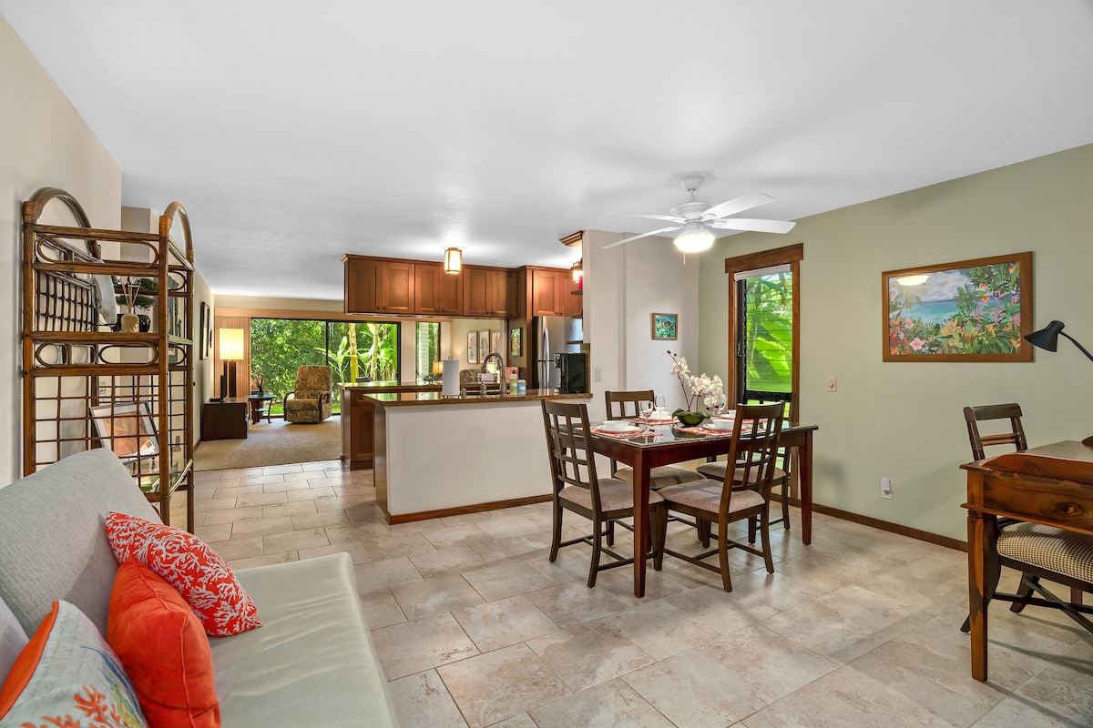 The open living space features a comfortable sofa with decorative pillows, a dining table for four, and a well-equipped kitchen in the background. Natural light fills the room through large sliding doors, providing a view of the lush surroundings.