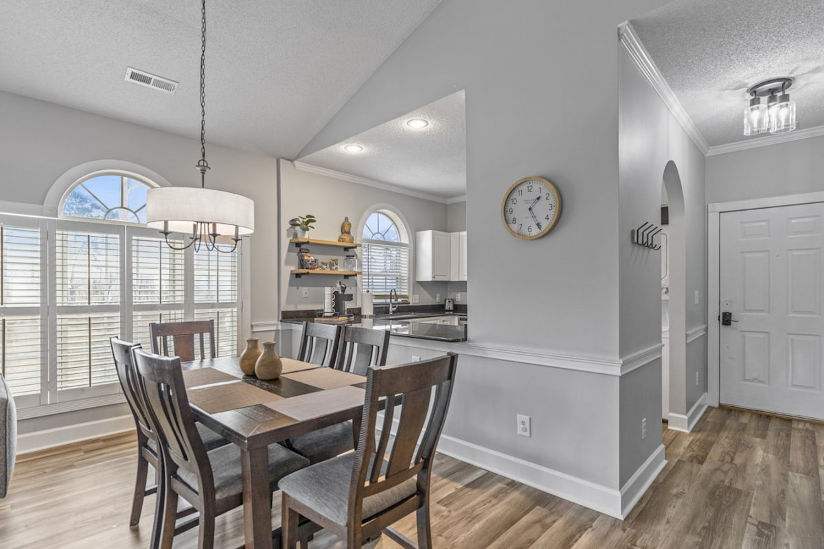 A dining area is featured with a wooden table and four chairs. Large windows allow natural light to fill the space, while a modern chandelier hangs overhead. The kitchen area is partially visible, showcasing sleek cabinetry and essential appliances.