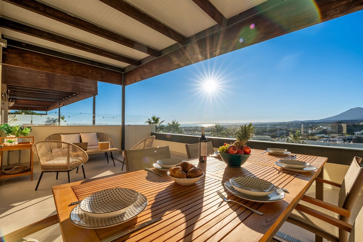 An outdoor terrace is furnished with a wooden dining table set for eight, featuring decorative plates and a bowl of fresh fruit. Woven chairs provide comfortable seating. The sun shines brightly in the sky, illuminating the views of the surrounding landscape and the sea in the distance.