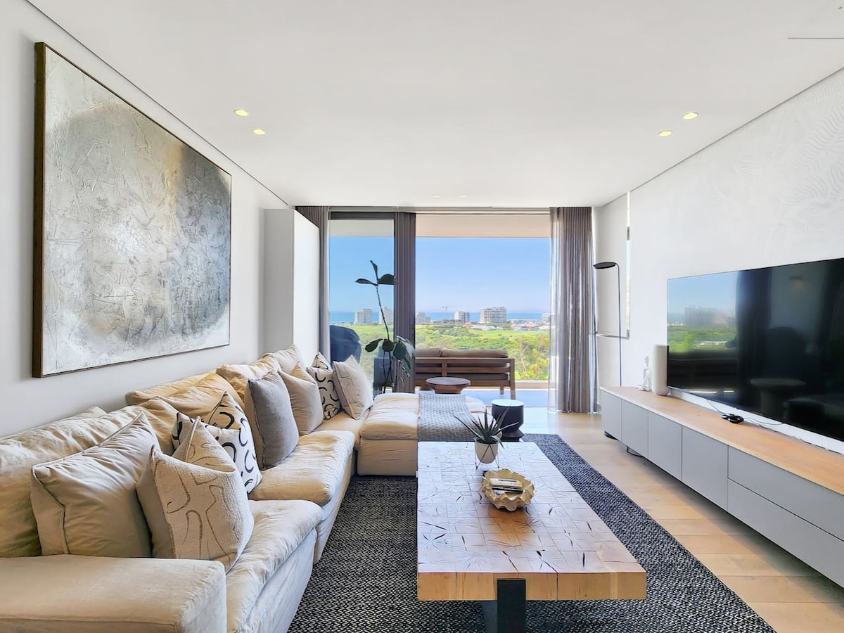 A spacious living area features a large sectional sofa adorned with neutral-toned cushions, positioned opposite a sleek television unit. A wooden coffee table rests in the center, and light streams in through expansive sliding glass doors, revealing views of the ocean and lush surroundings.