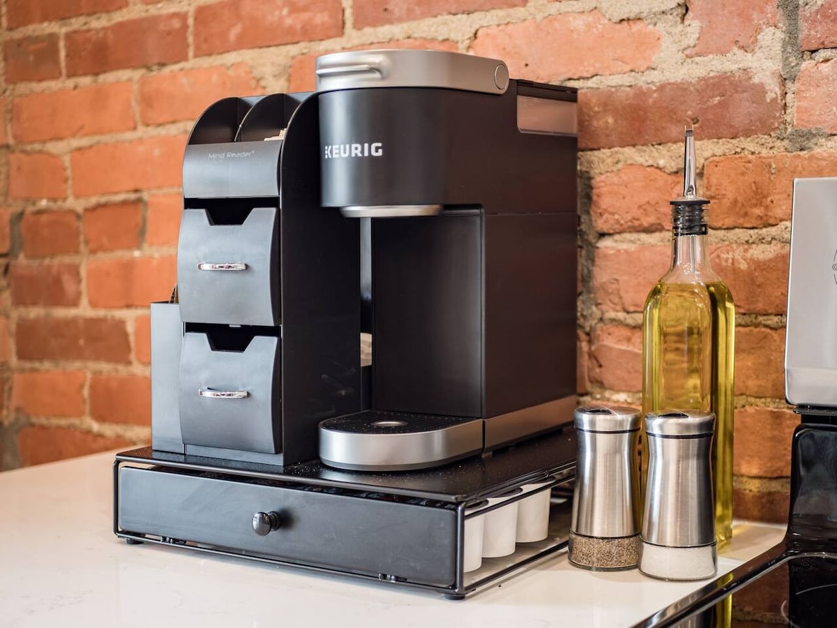A Keurig coffee maker is prominently displayed on a countertop against a backdrop of exposed brick. Adjacent items include a stackable pod holder for coffee pods, a glass bottle of olive oil, and a set of salt and pepper shakers in metallic containers.