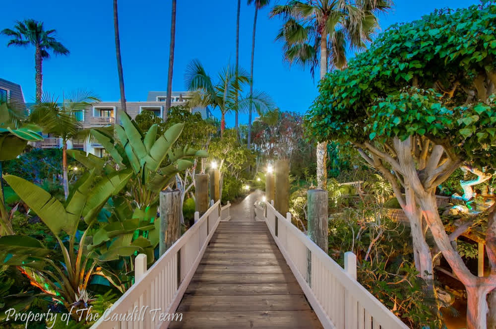 A wooden walkway leads through lush tropical landscaping under a dusk sky. Palm trees and vibrant plants frame the path, while soft lighting enhances the serene atmosphere.