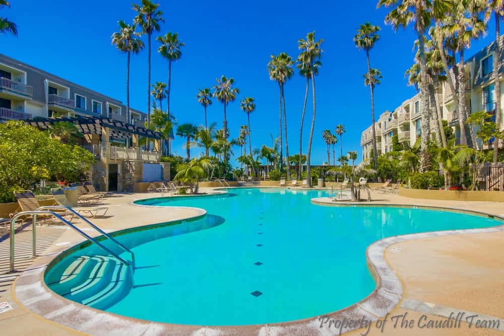 A large, inviting pool is surrounded by palm trees and tropical landscaping. Lounge chairs are arranged around the water's edge, inviting relaxation. The clear blue sky reflects on the pool's surface, contributing to a serene atmosphere within the outdoor area.