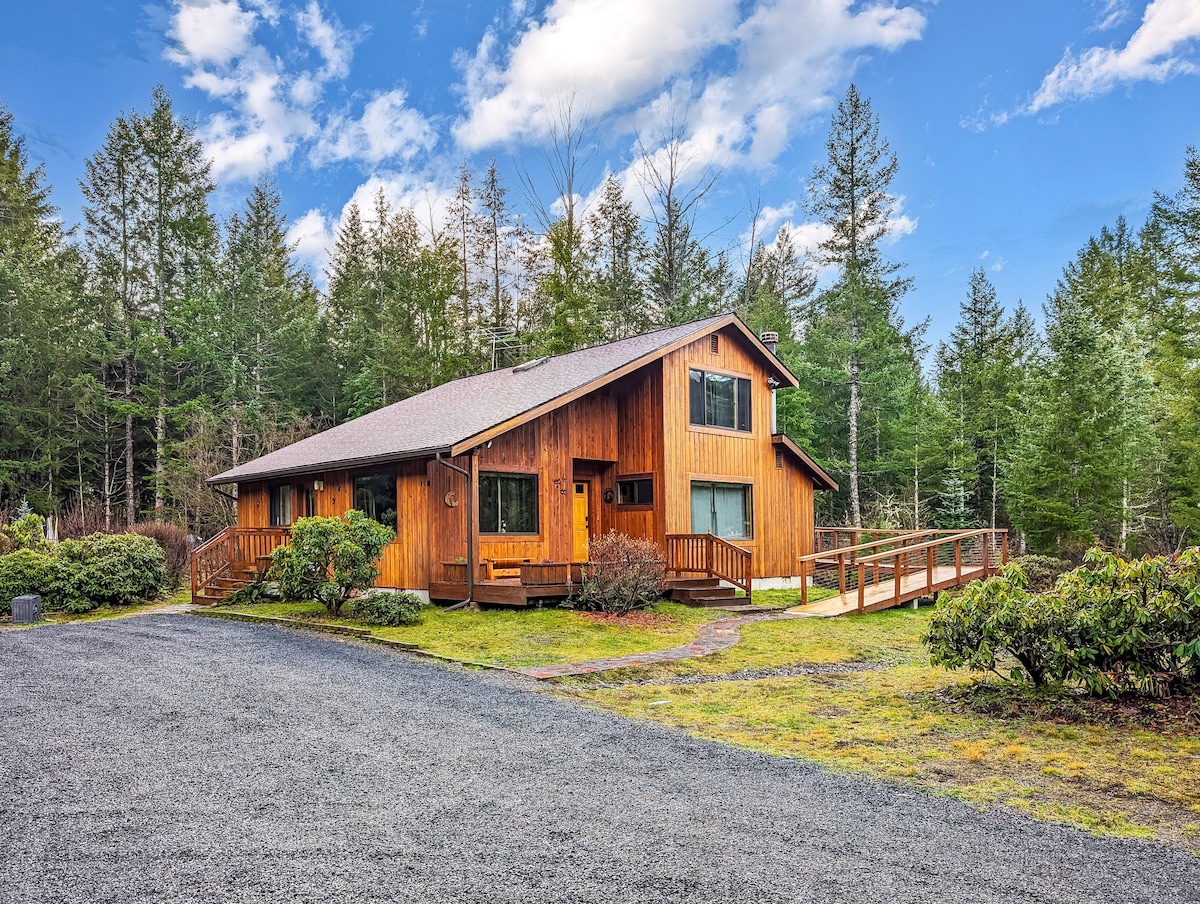 A two-story wooden house is set amidst lush greenery, featuring a welcoming front porch and wheelchair-accessible ramp. Surrounding shrubs and a gravel driveway contribute to the natural ambiance, while tall trees frame the scene, highlighting the peaceful outdoor setting.