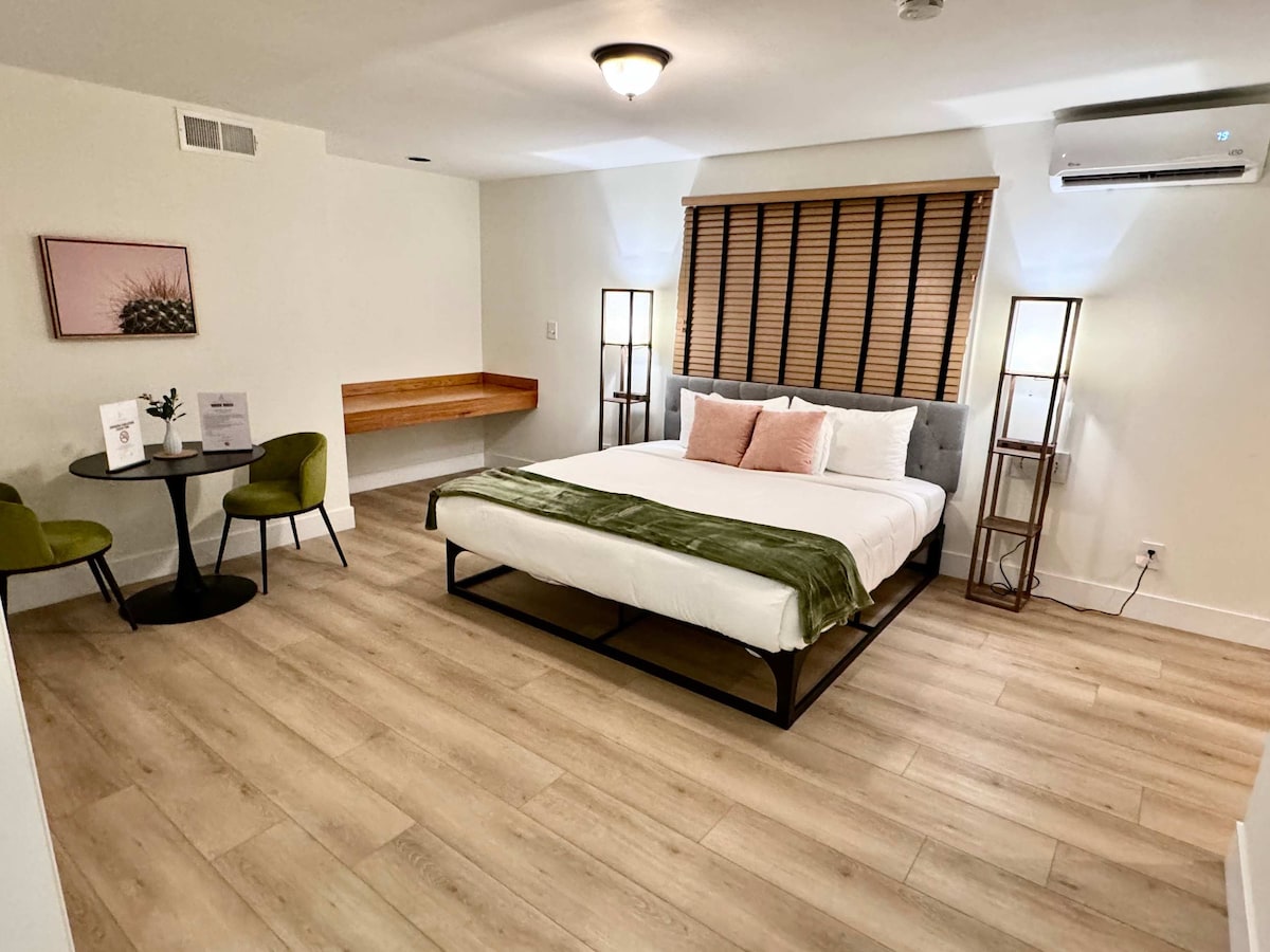 A comfortable room features a spacious bed with a light green throw blanket and decorative pillows. Two modern lamps stand beside the bed, while a small dining area with two chairs and a table is situated by the window. Light-colored wooden flooring enhances the space.