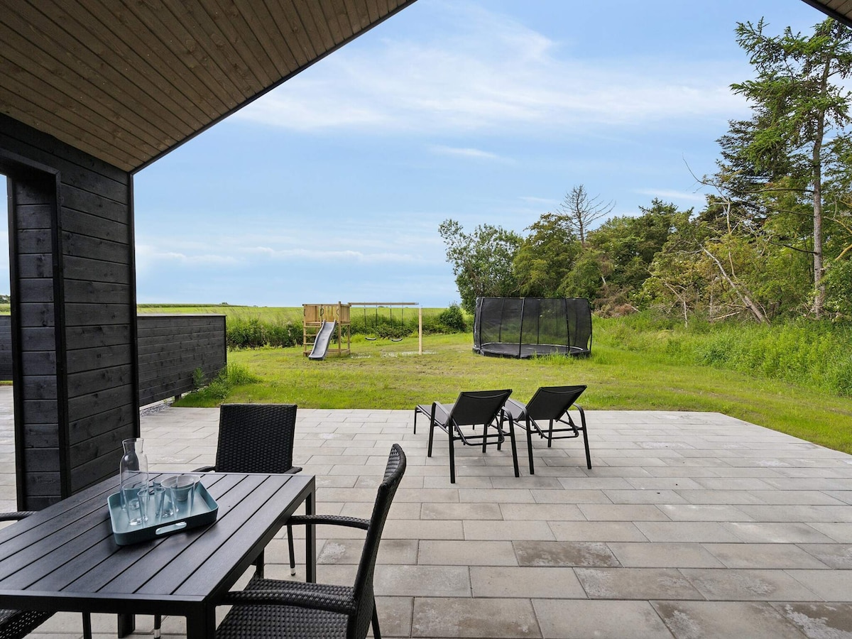 An outdoor patio is positioned with stone flooring, featuring a table and chairs alongside two lounge chairs. A grassy area is visible, equipped with a playground structure and a trampoline, framed by natural greenery and a distant view of open land.