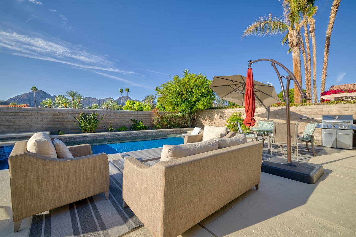 A tranquil outdoor area features a refreshing pool bordered by lush greenery and palm trees. Comfortable beige couches sit on a light-colored patio, while a shaded dining space is equipped with a grill and seating. Mountains are visible in the background under a clear blue sky.