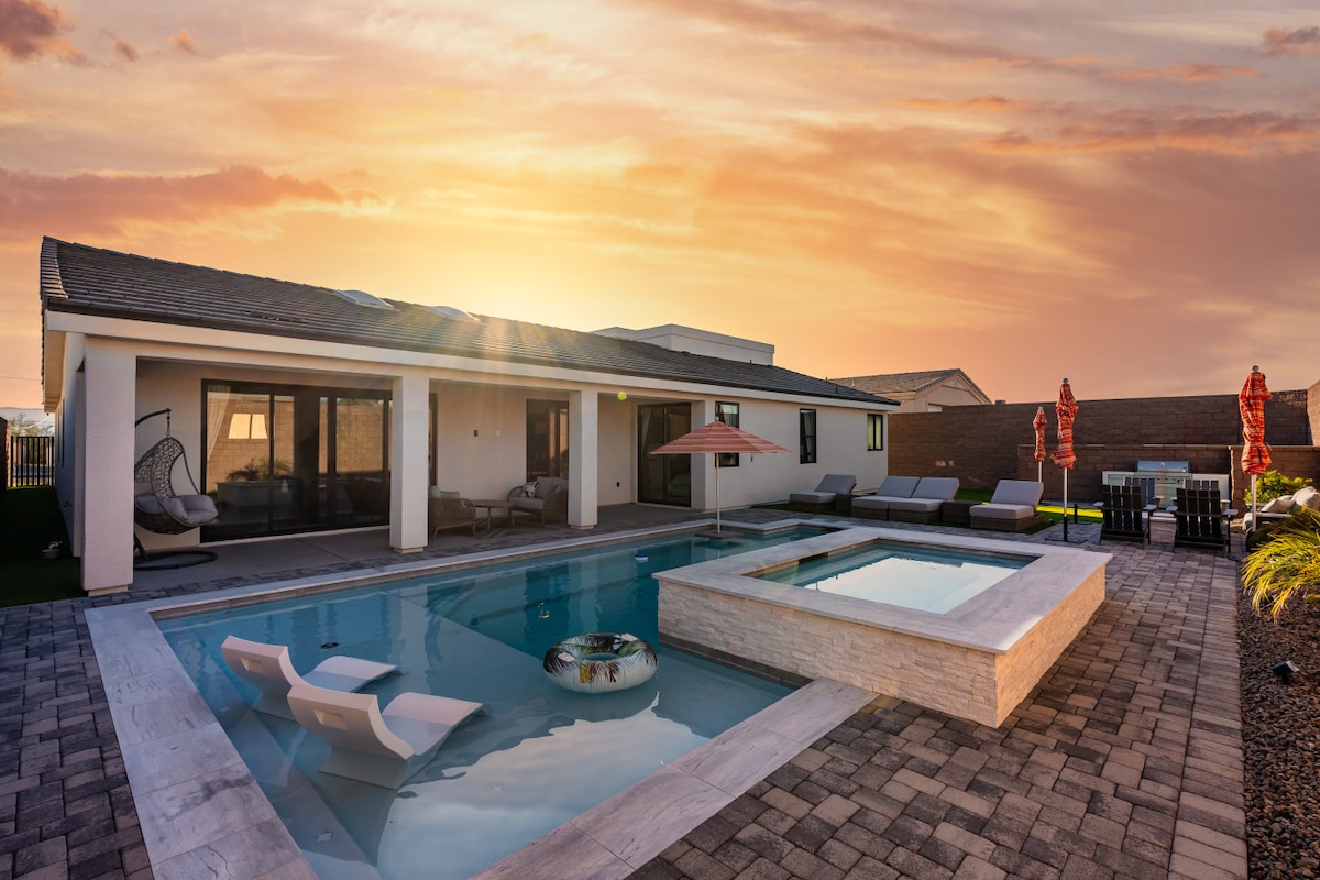 The expansive backyard features a sleek swimming pool with loungers and an integrated spa. A shaded seating area is arranged for relaxation, complemented by vibrant red umbrellas. The home’s modern architecture is visible, framed by a stunning sunset casting warm tones across the landscape.