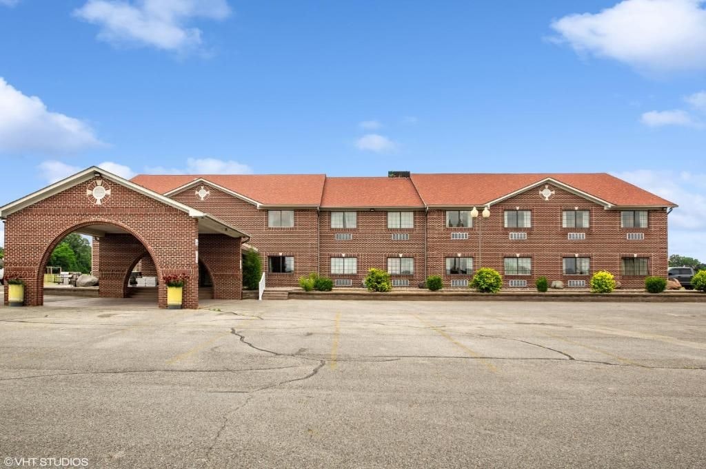 The exterior of the hotel features a classic brick facade complemented by a gabled entrance. Multiple windows can be seen on the upper level, while an ample parking area is present in the foreground. Lush landscaping adds subtle color around the entrance.