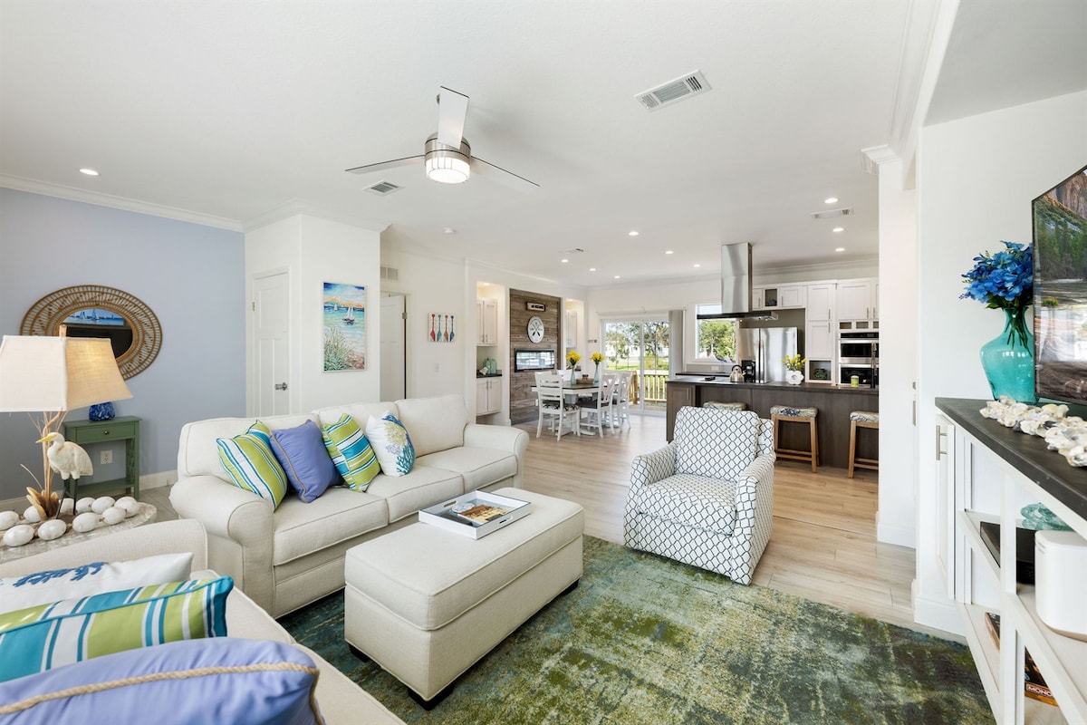 A spacious living area features comfortable seating with soft, neutral tones and colorful accent pillows. A large area rug anchors the space, while natural light floods in through large windows, creating a bright ambiance that connects to the open kitchen beyond.