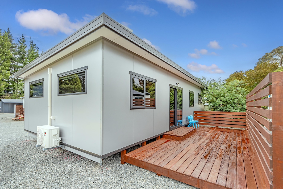 The exterior of the accommodation features a modern design with large windows allowing ample natural light. A wooden deck is accessible from the entrance, complemented by a small blue chair. The surrounding area includes gravel and greenery, providing a serene outdoor setting.