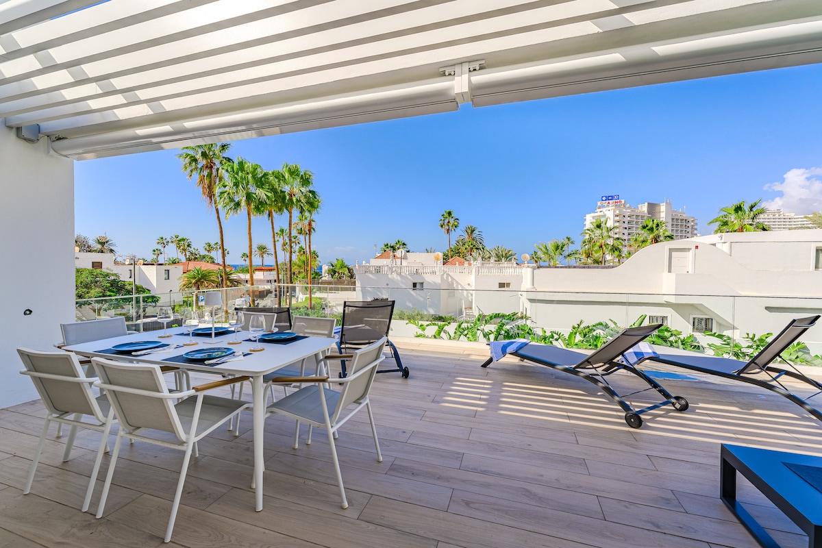 An outdoor terrace features a dining table set for six, complemented by loungers for relaxation. Tropical greenery frames the view, enhancing a serene atmosphere. Light wooden flooring extends throughout the space under a shaded awning, providing comfort for outdoor dining and lounging.