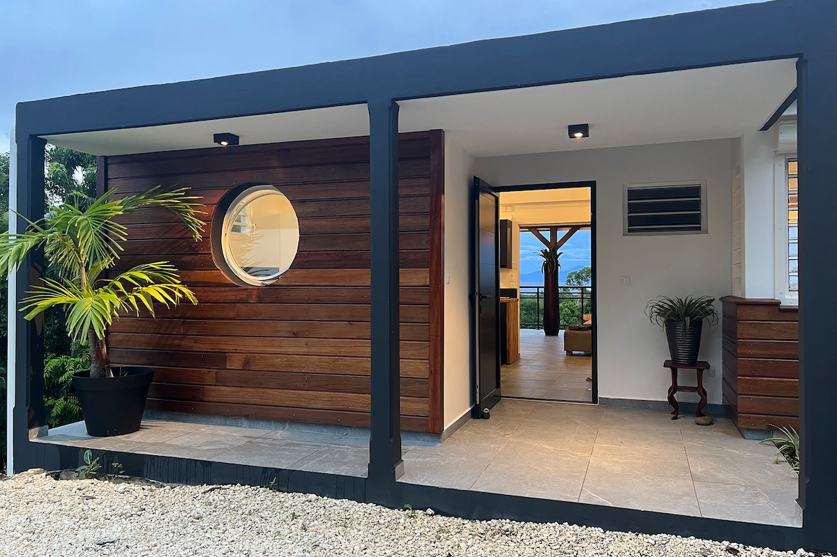 An inviting entrance is framed by a sleek black structure featuring warm wooden accents. A round window enhances the aesthetic, while a potted palm adds a touch of greenery. Visible through the open door, the well-lit living space extends into a serene indoor-outdoor flow.