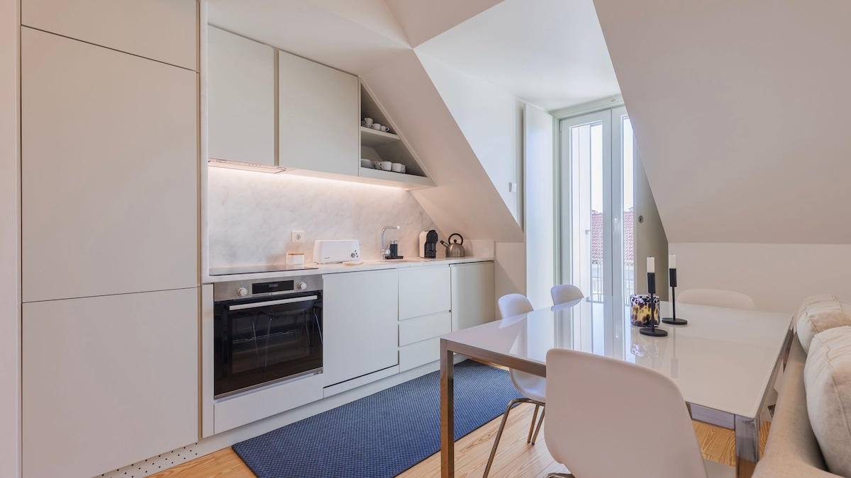 A modern kitchenette features sleek cabinetry made of light-colored materials, complemented by a marble countertop. A dining table with white chairs is situated nearby. Large windows offer ample natural light, while a gray area rug adds warmth to the space.