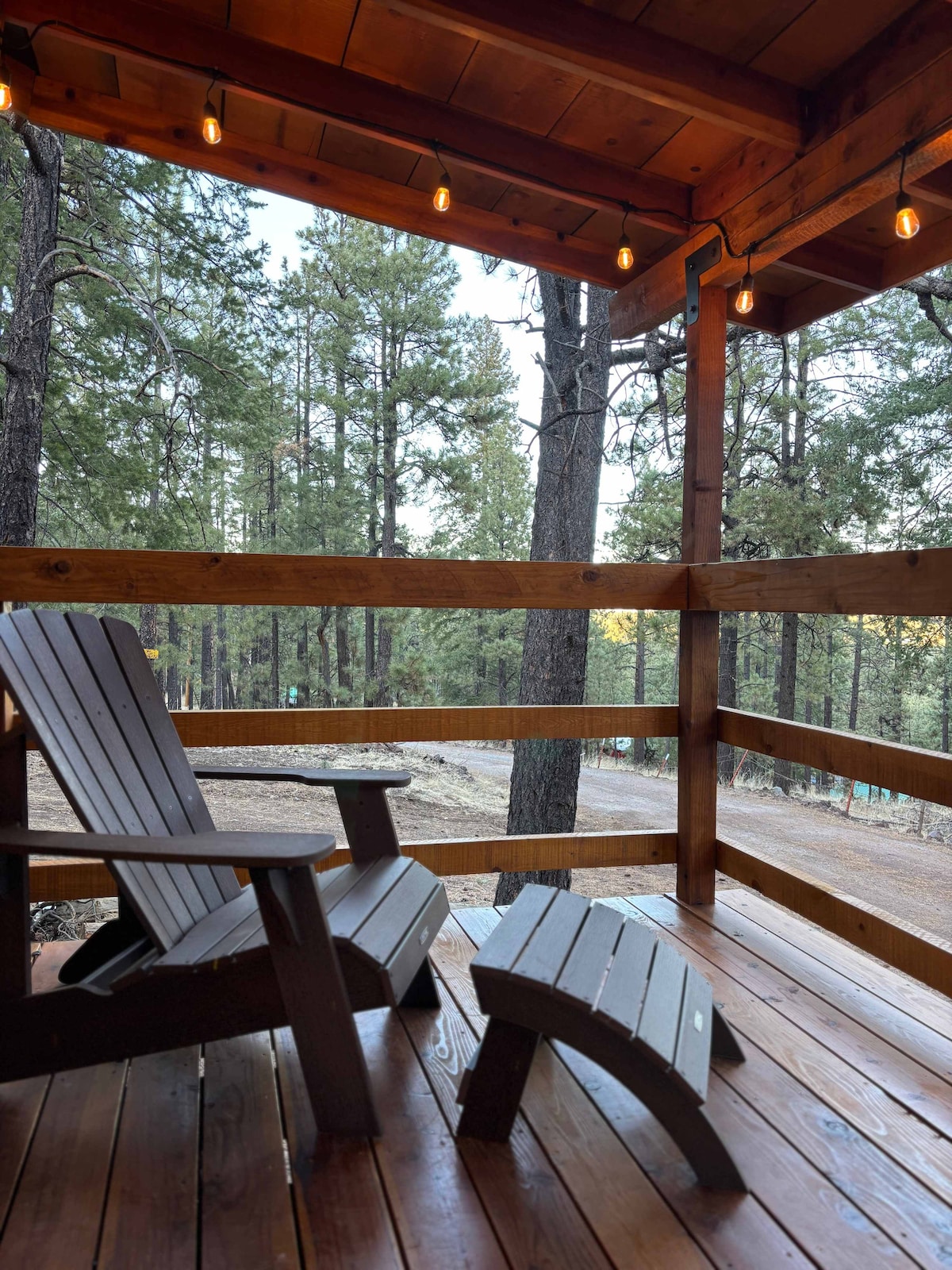 A wooden porch with a rustic feel features two Adirondack chairs and a small side table, surrounded by tall pine trees. Soft string lights hang above, enhancing the natural ambiance of the outdoor space.