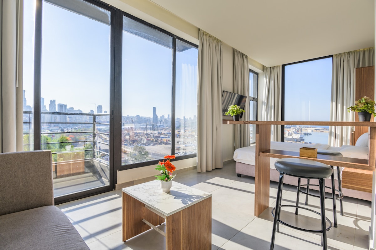 A bright and airy interior features large floor-to-ceiling windows that frame stunning views of the city and sea. A cozy seating area includes a plush sofa and modern coffee table with a small potted plant. The dining counter adds functionality, providing a space for meals with a couple of stylish stools nearby.