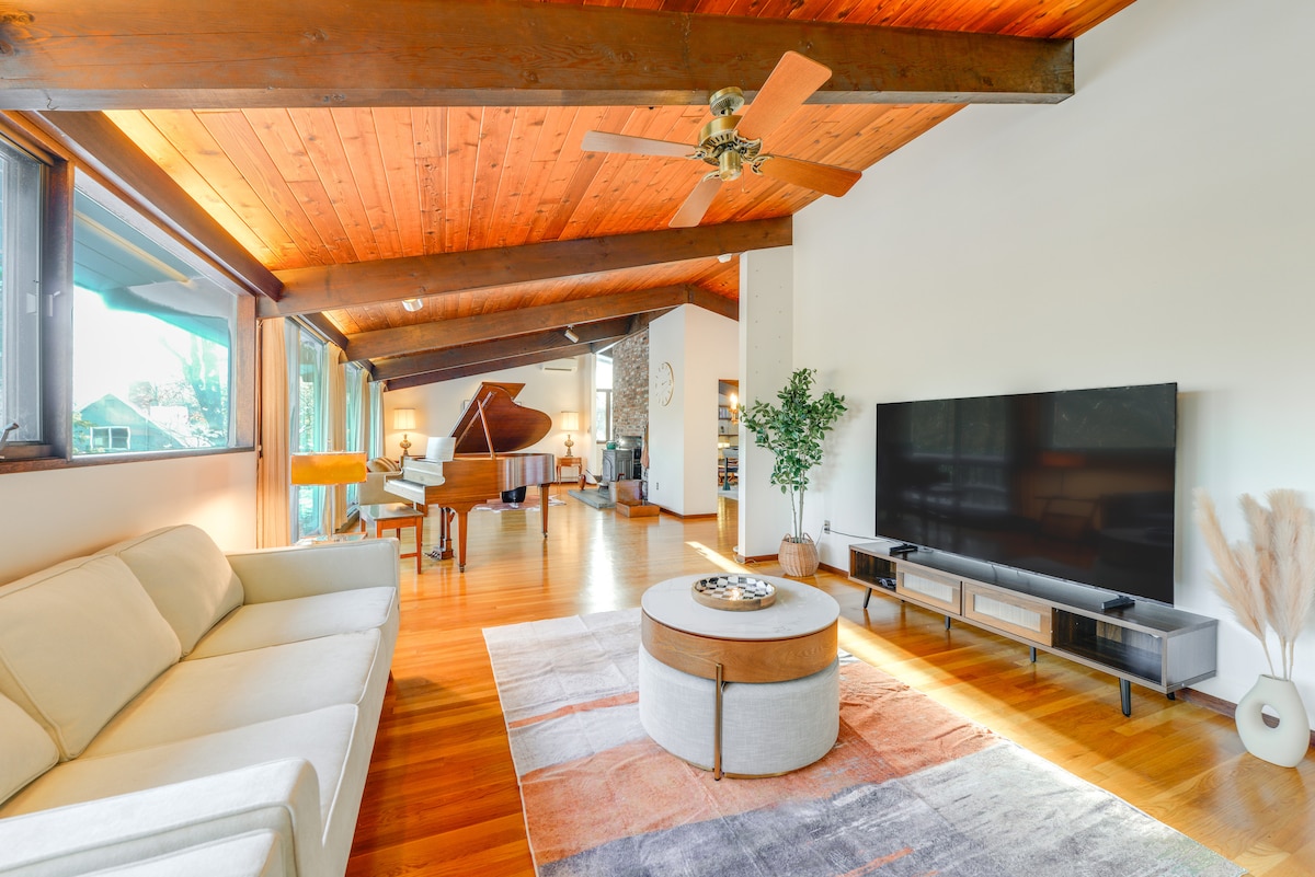 The spacious living area features vaulted wooden ceilings and large windows, allowing natural light to fill the space. A beige sectional sofa is positioned on a colorful area rug, with a coffee table in the center. A baby grand piano is seen in the background.