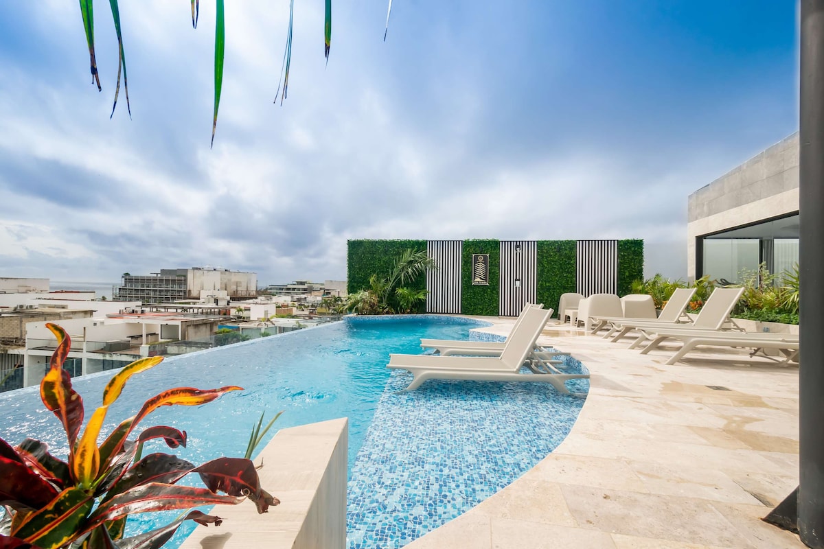 The shared rooftop pool features an infinity edge, presenting panoramic views of the ocean and surrounding cityscape. Sun loungers are positioned along the deck, with vibrant plants and greenery enhancing the outdoor space.
