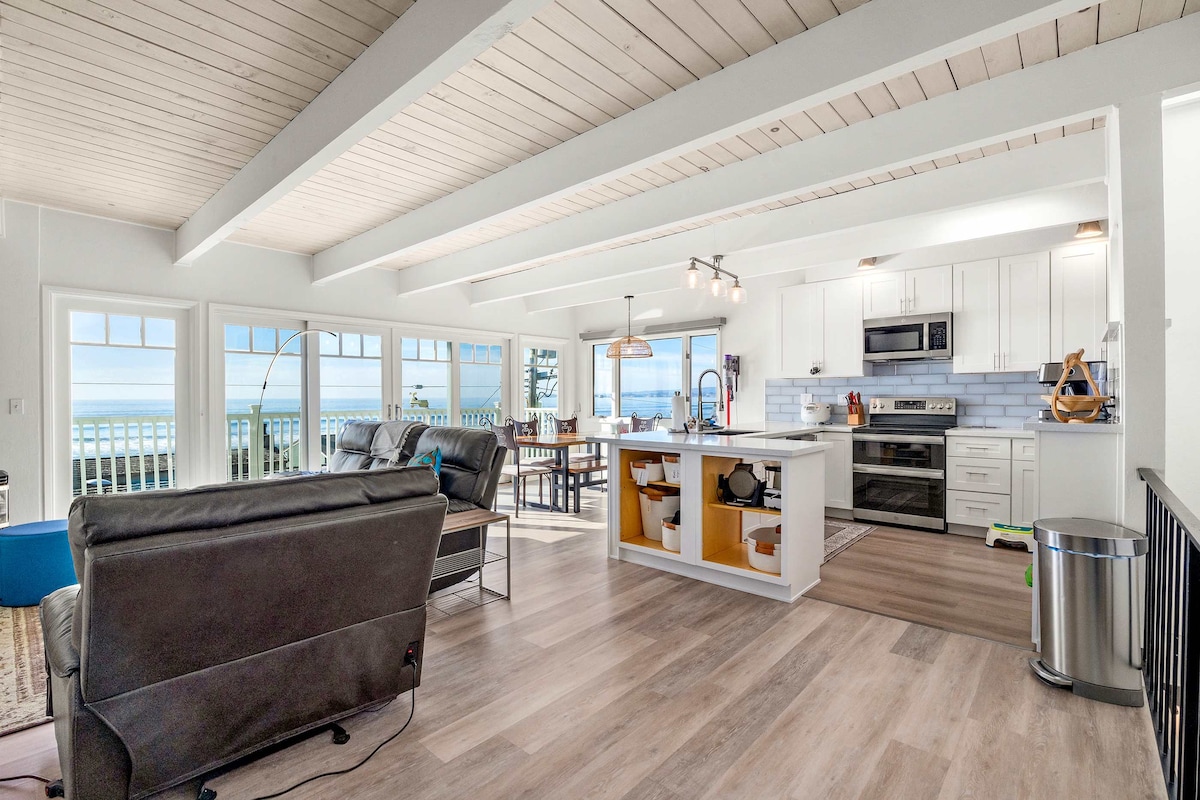 An open-concept living space features a modern kitchen with stainless steel appliances and ample counter space. Light-colored walls and exposed beams create a bright atmosphere. Large windows provide views of the ocean, while a comfortable seating area accommodates relaxation. The dining area is positioned nearby.