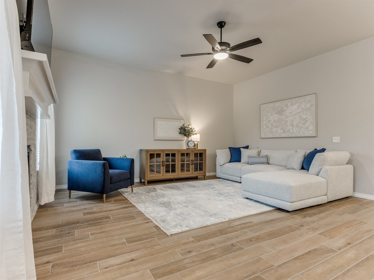 A spacious living room is highlighted by a large sectional sofa and a cozy accent chair. A wooden media console sits against the wall, and a light area rug covers the floor. Large windows allow natural light to fill the room, complemented by a ceiling fan.