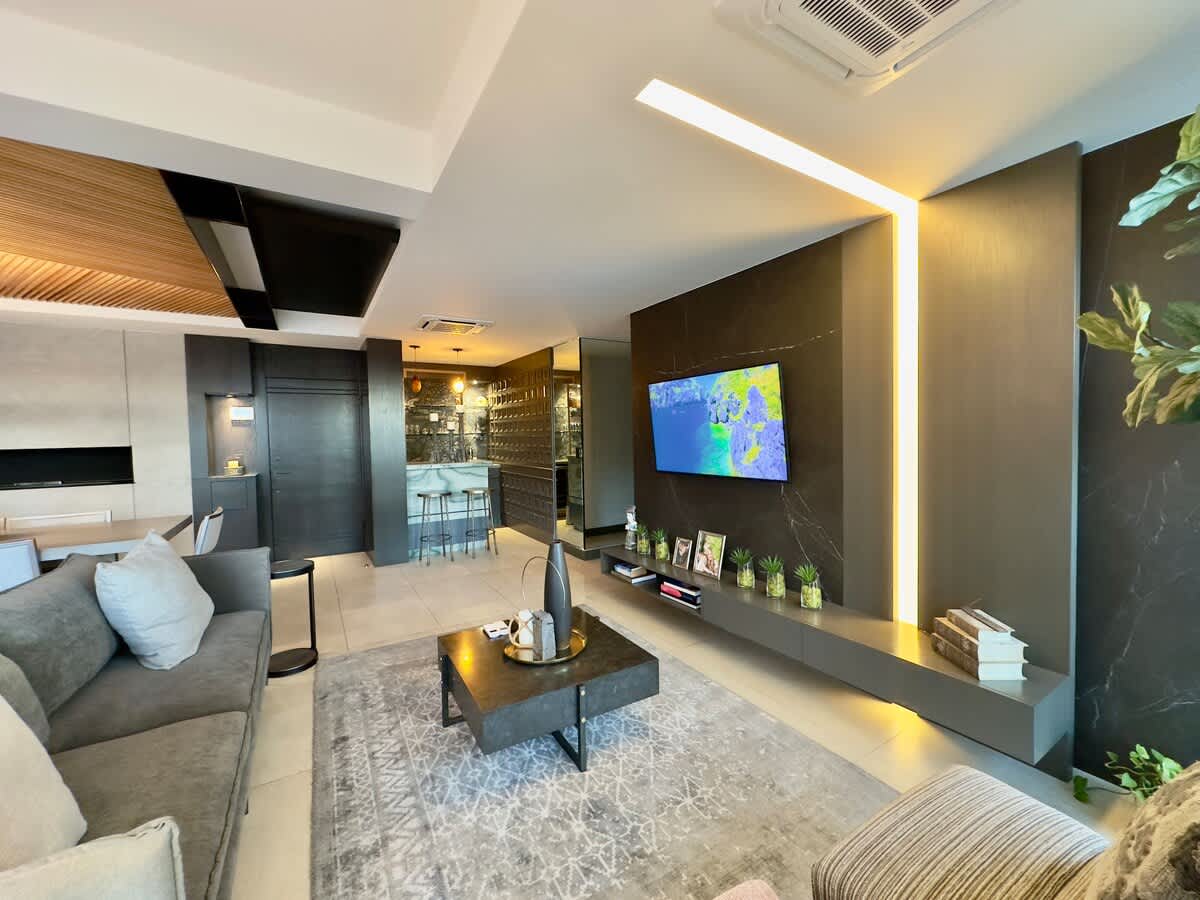 A modern living area is presented with a light grey sofa and a large, dark coffee table. A flat-screen television is mounted on the wall, surrounded by decorative plants. A stylish kitchenette is visible in the background, enhancing the open concept design.