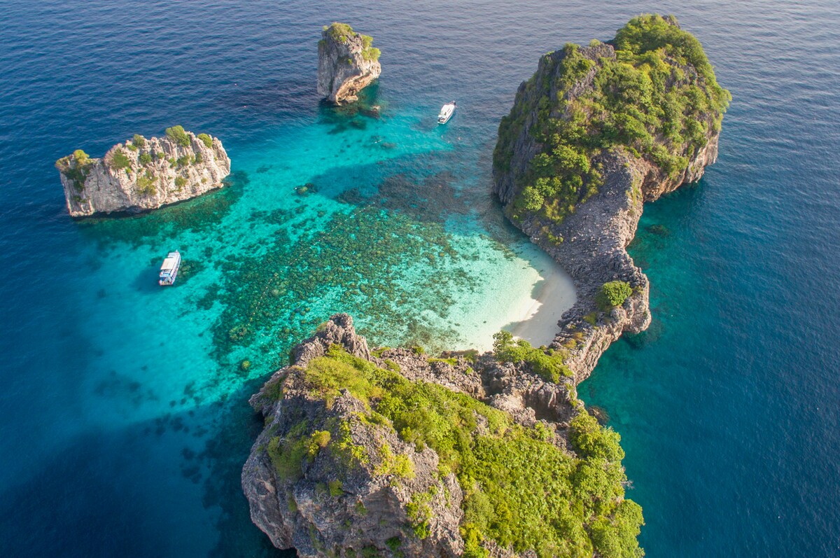Aerial view of a secluded cove surrounded by rocky islands and shallow turquoise waters. Lush greenery blankets the rocks, while two boats are visible in the distance. The white sandy beach is nestled between the natural formations, highlighting the tranquil beauty of the landscape.