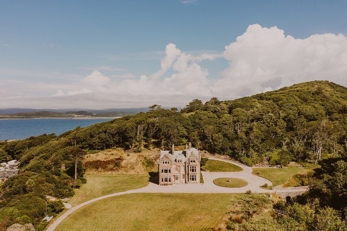 An aerial view captures a grand two-story house nestled amidst lush greenery, with the sea visible beyond. Elevated on a hillside, the property is surrounded by expansive landscaped grounds featuring a sweeping driveway and circular lawn.