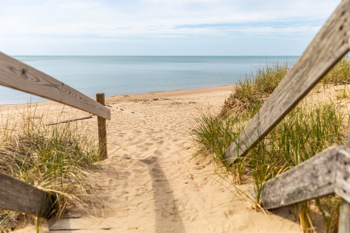 A sandy pathway leads through tall grass to the calm waters of Lake Michigan. Wooden steps descend towards the beach, with a serene horizon visible in the background under a clear sky.