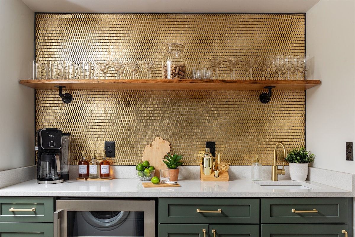 A modern kitchen area features a textured gold backsplash paired with wooden shelving. Glassware and decorative items are neatly arranged above a countertop with a coffee maker, citrus fruits, and a gold barware set. A stainless steel sink adds a sleek touch to the design.