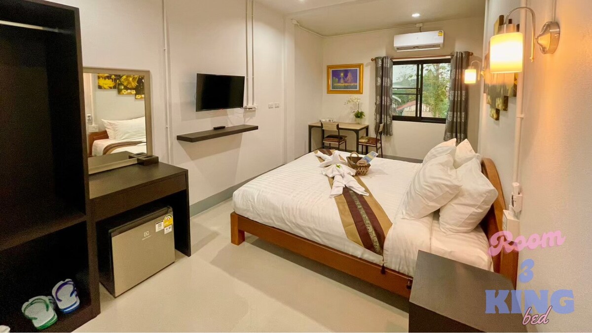 The room features a spacious bed adorned with decorative pillows. A flat-screen television is mounted on the wall opposite the bed. A small table is set for dining, while a window provides natural light, framed by curtains. Additional furniture includes a shelf and a mini-fridge.