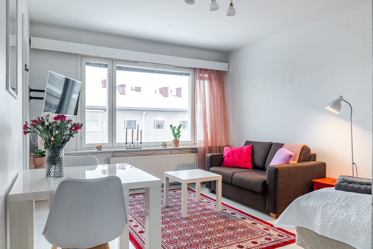 A well-lit living area features a comfortable dark sofa with colorful cushions, complemented by a simple white coffee table. A fresh flower arrangement occupies the table, while a decorative rug adds warmth. Large windows with light curtains allow natural light to enter the space.