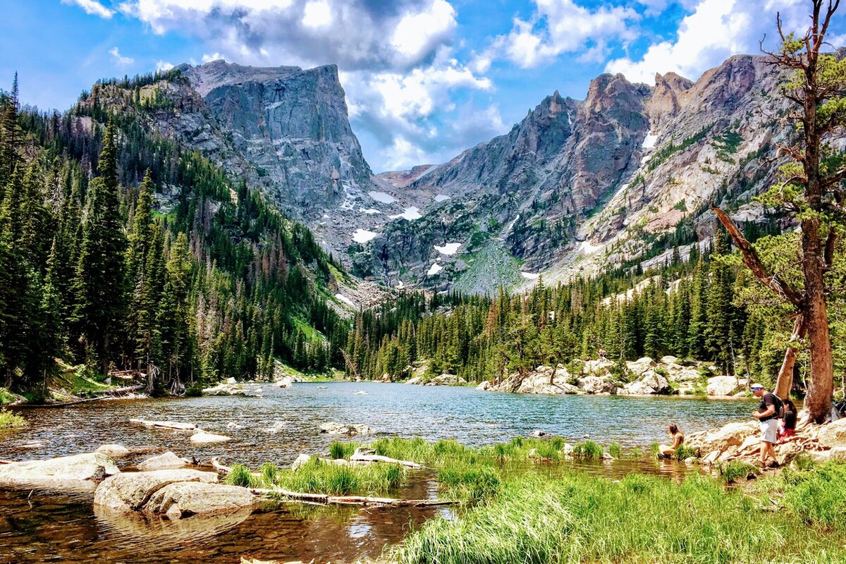 A serene mountain landscape is depicted, featuring a clear lake surrounded by lush green trees and rocky terrain. Majestic mountains rise in the background, partially covered in snow. Two individuals can be seen wading in the shallow water, enjoying the peaceful environment.