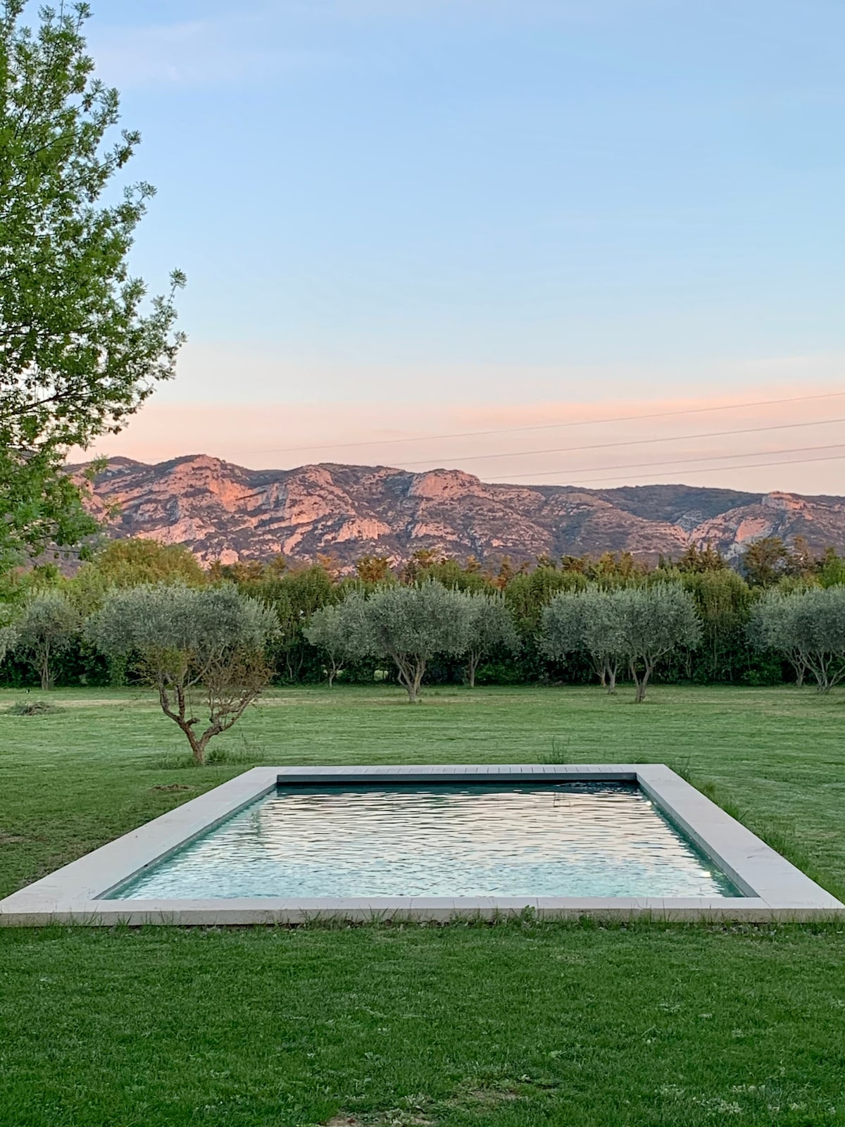 A serene view of a private pool reflects the surrounding landscape, framed by neatly trimmed grass. Olive trees are visible in the distance, with majestic mountains bathed in soft evening light, creating a tranquil atmosphere.