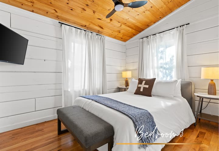 Guest bedroom offers a serene retreat with a queen-sized bed, crisp white linens, and a warm wood ceiling. Combining comfort and rustic charm.