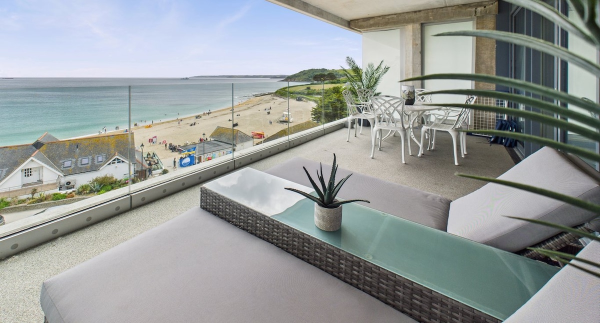 A spacious balcony provides stunning sea views and direct beach access. Relaxing lounge chairs are placed near a glass table, accompanied by a small plant centerpiece. The beach is visible in the distance, with sand and waves creating a serene coastal atmosphere.