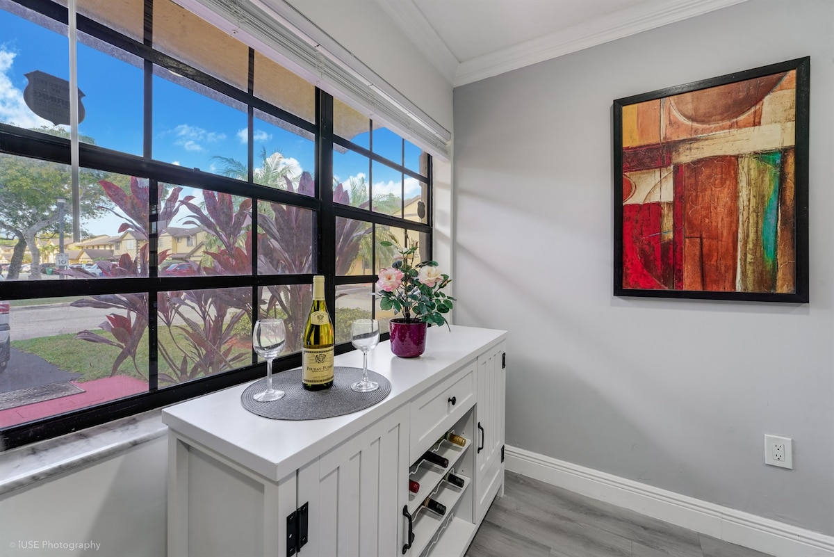 A neatly arranged console table displays a bottle of champagne and two glasses. Fresh flowers add a touch of color, while a vibrant artwork piece hangs on the wall. Large windows offer natural light and a view of the outdoors.