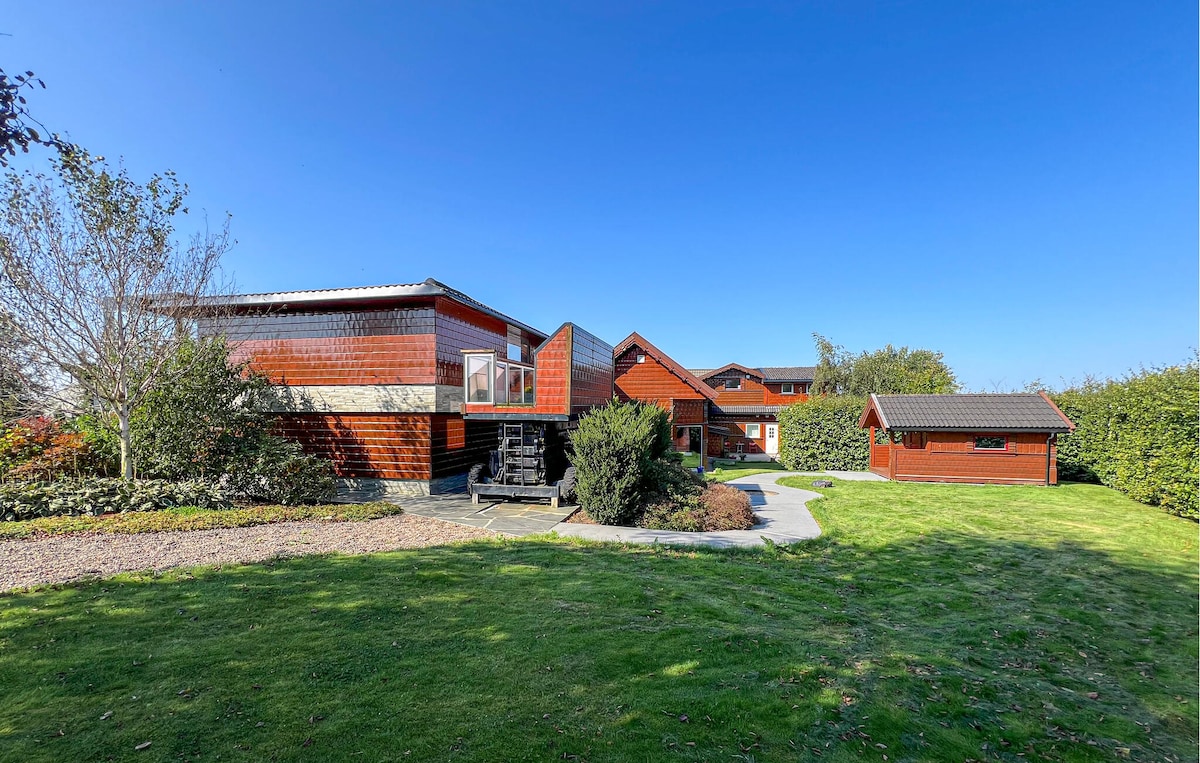 The holiday home is set amidst a landscaped garden with lush green grass and well-maintained shrubbery. The exterior features a mix of wooden textures in warm tones. Clear blue skies frame the charming buildings, including a separate storage shed visible in the background.