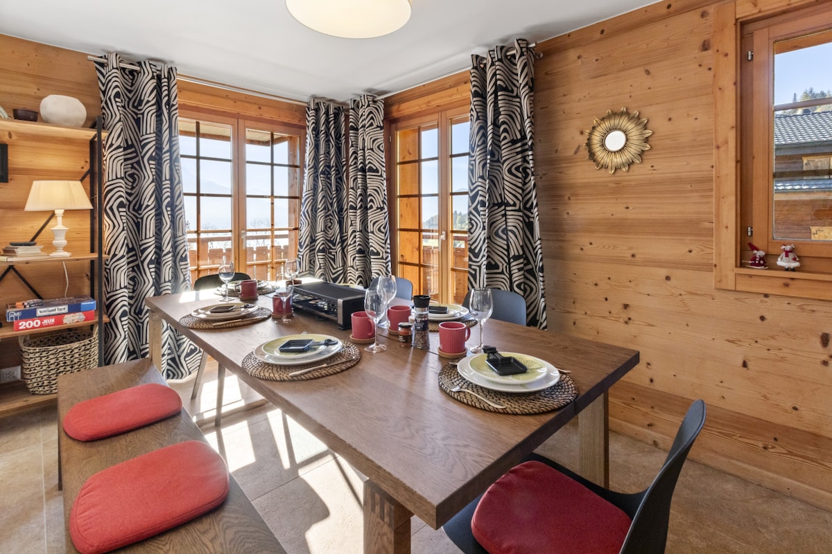 A dining area features a large wooden table set with plates and glassware. Four chairs surround the table, complemented by padded benches. Natural light enters through multiple windows, while patterned curtains add texture to the wooden walls.