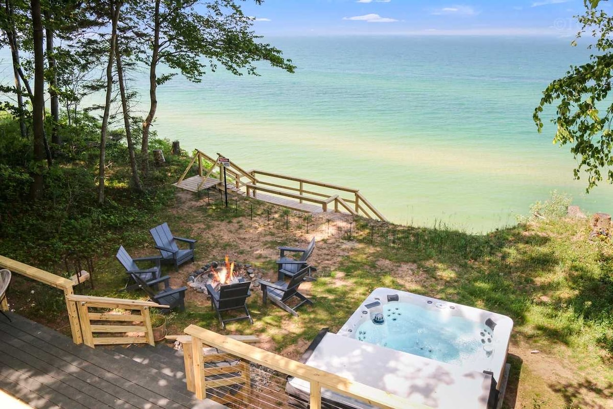 A serene outdoor space featuring a hot tub adjacent to a firepit surrounded by lounge chairs. A wooden deck extends toward the lake, where stairs lead down to the water's edge. The view captures the tranquil blue hues of Lake Michigan and the greenery of the surrounding trees.