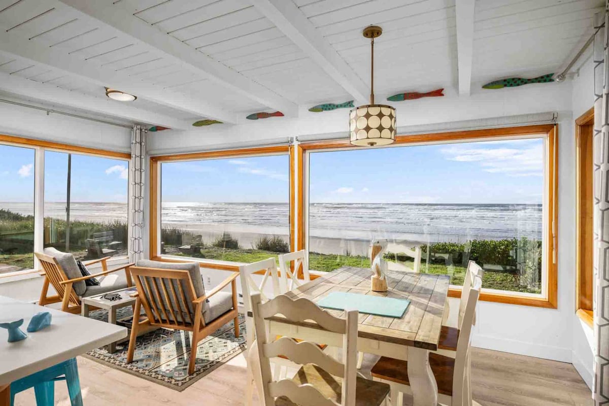 A bright dining and sitting area features large picture windows offering panoramic views of the ocean. Comfortable seating surrounds a rustic dining table, while natural light floods the space. Coastal décor accents the room, enhancing the relaxed beach ambiance.