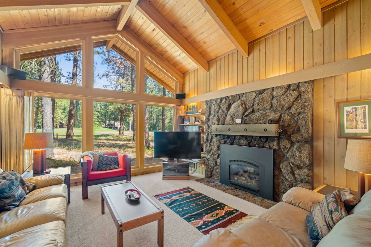 The main living area is adorned with natural wood features, including vaulted ceilings and exposed beams. Large windows offer views of the golf course, while a stone fireplace serves as a focal point. Comfortable seating is arranged around a coffee table, with decorative elements enhancing the space.