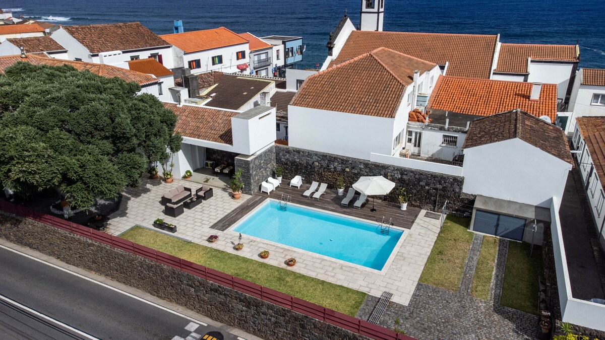 An aerial view showcases a rectangular swimming pool framed by a well-maintained patio area, featuring sun loungers and a shaded seating arrangement. Surrounding gardens with potted plants provide greenery, while the coastline is visible in the background, complemented by distinct red-tiled roofs.