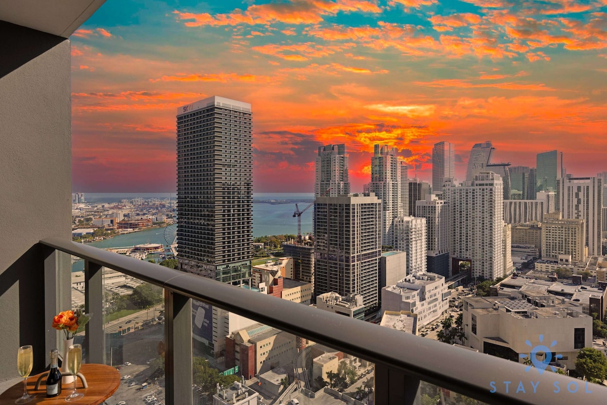 A balcony view showcases a stunning cityscape at sunset, with vibrant oranges and reds illuminating the skyline. Tall buildings and water can be seen in the distance, creating a serene and scenic backdrop for relaxation.