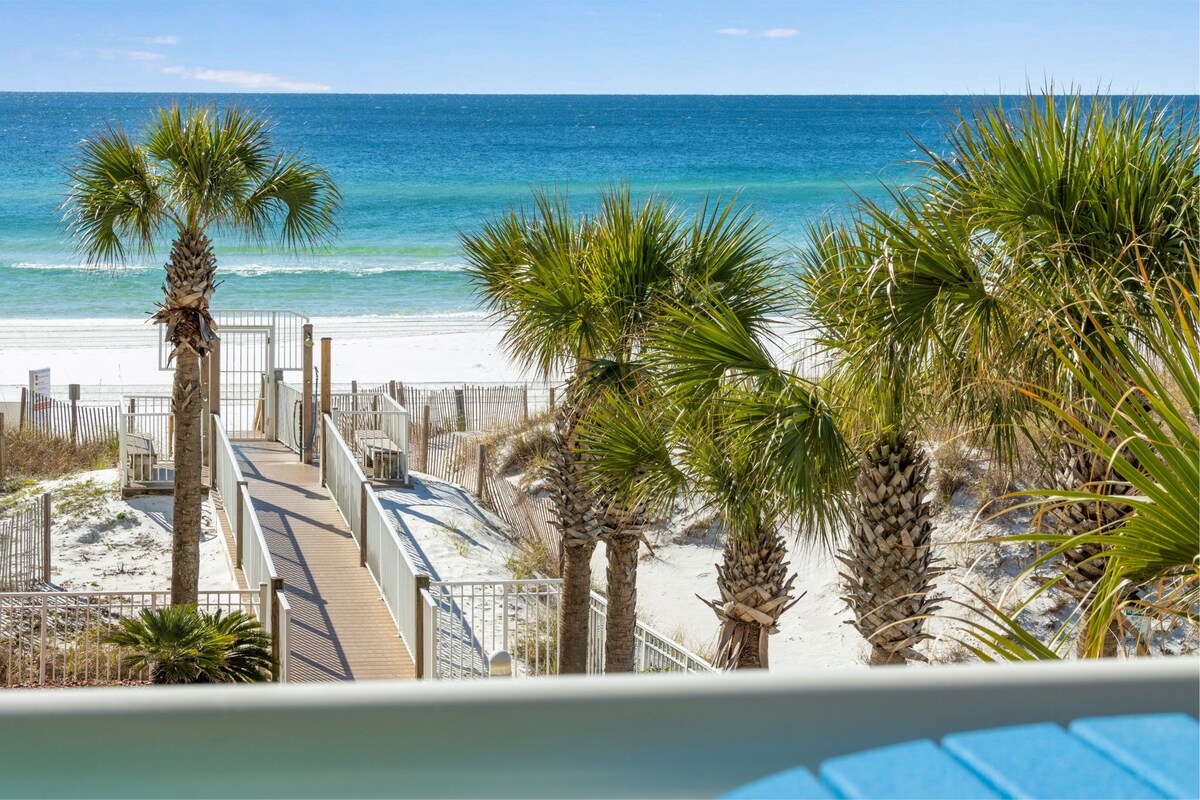 A view of the private beach access pathway lined with palm trees. The sandy beach and crystal-clear waters of the Gulf are visible in the distance under a bright blue sky, providing a serene coastal atmosphere.