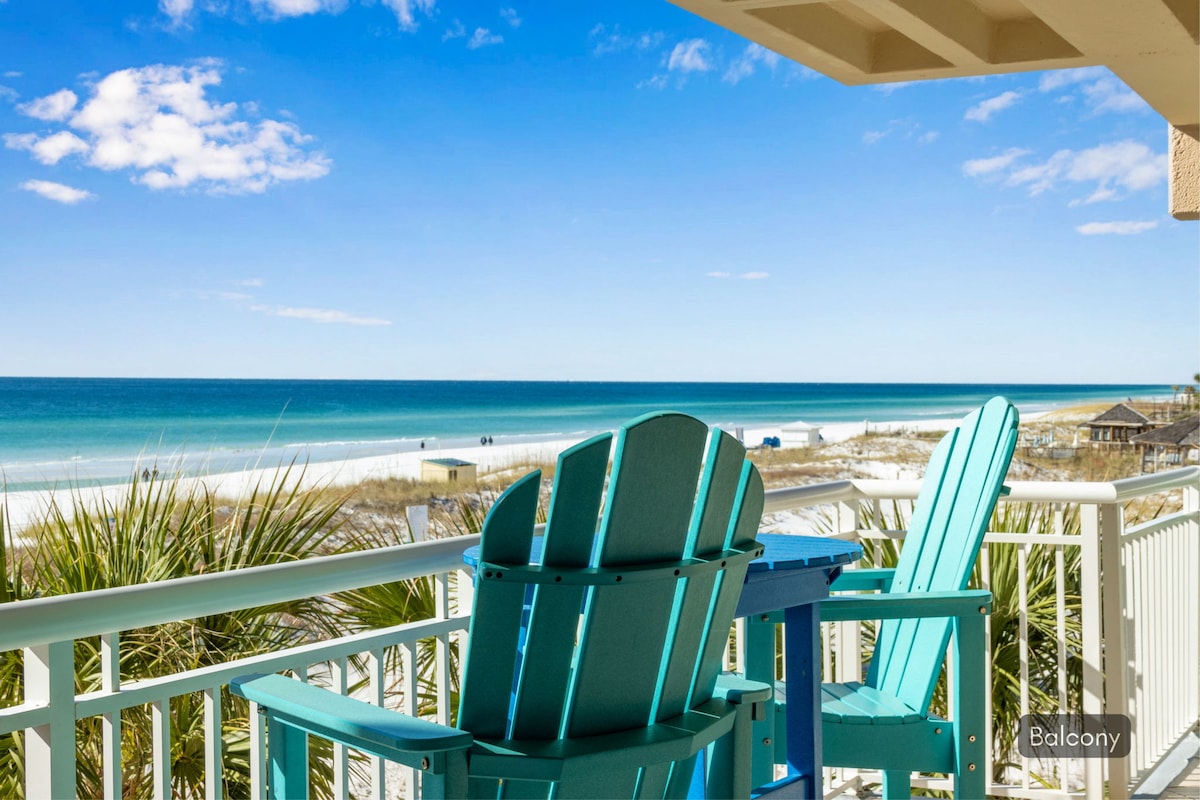 The image captures a balcony view overlooking the beach, featuring two turquoise Adirondack chairs. The expansive horizon showcases the tranquil blue waters of the Gulf under a bright sky with scattered clouds, framed by lush greenery and sandy beach paths.