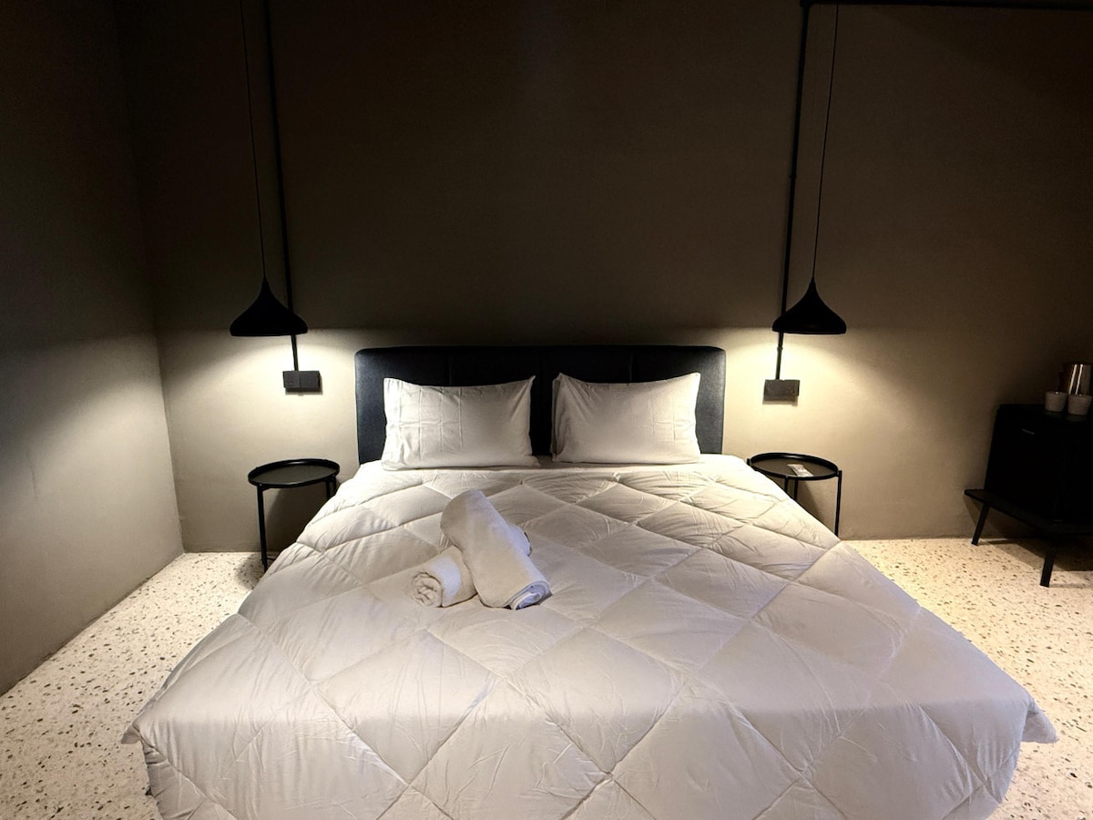 A spacious queen bed is centered in the room, illuminated by two pendant lights on either side. Crisp white bedding is neatly arranged, complemented by two decorative pillows. The floor features a textured design, while minimalistic nightstands are positioned at each side of the bed.